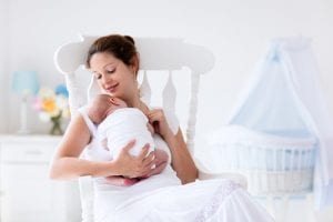 Woman and new born boy relax in a white bedroom with rocking chair and blue crib