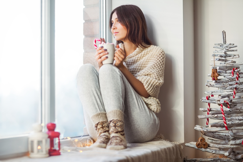 Happy beautiful woman drinking hot coffee sitting on window sill in christmas decorated home-img-blog